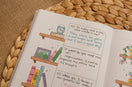 Open A5 guided journal featuring a bookshelf-themed weekly reflection spread with colorful illustrated books, handwritten journaling prompts, and cozy neutral styling on a woven placemat.