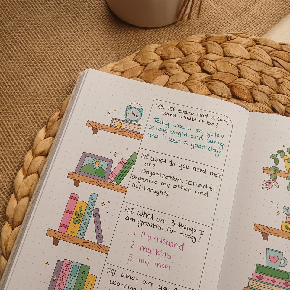 Open A5 guided journal featuring a bookshelf-themed weekly reflection spread with colorful illustrated books, handwritten journaling prompts, and cozy neutral styling on a woven placemat.