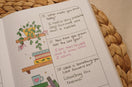 Close-up of an open guided journal with bookshelf illustrations, handwritten weekly reflection prompts, and colorful ink on a dotted page, styled on a natural woven mat with warm neutral décor.