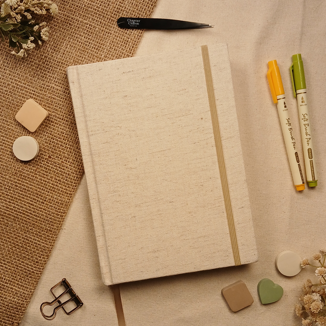 Minimalist linen Booked Journal with cream fabric cover, elastic closure, and ribbon bookmark styled with planner pens, tweezers, and neutral desk accessories on a linen background.