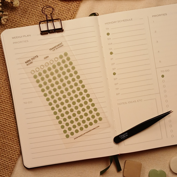 Transparent matte round planner dot stickers in Fern green displayed on an open weekly planner with Chapter & Charm tweezers on a warm neutral linen background.