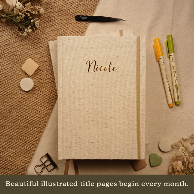 Personalized Booked Journal featuring the name "Nicole" engraved on the linen cover, shown with planner accessories. Text on image: "Beautiful illustrated title pages begin every month."