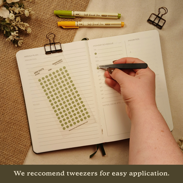 Hand using Chapter & Charm tweezers to place a Fern green transparent round planner sticker in an open planner. The image includes the text, "We recommend tweezers for easy application."
