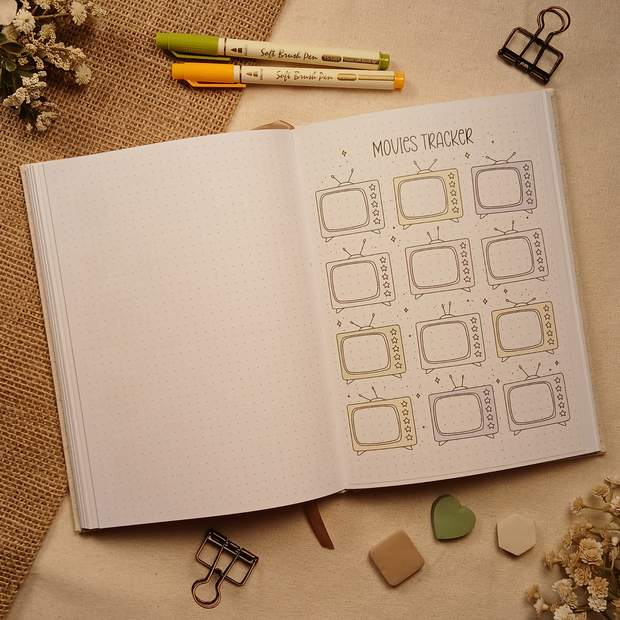 Open A5 dotted journal featuring a color Movies Tracker stick-in page with retro television illustrations accented in soft earth tones for recording favorite films and movie ratings. Styled with brush pens, binder clips, dried florals, burlap, and neutral sticky notes.
