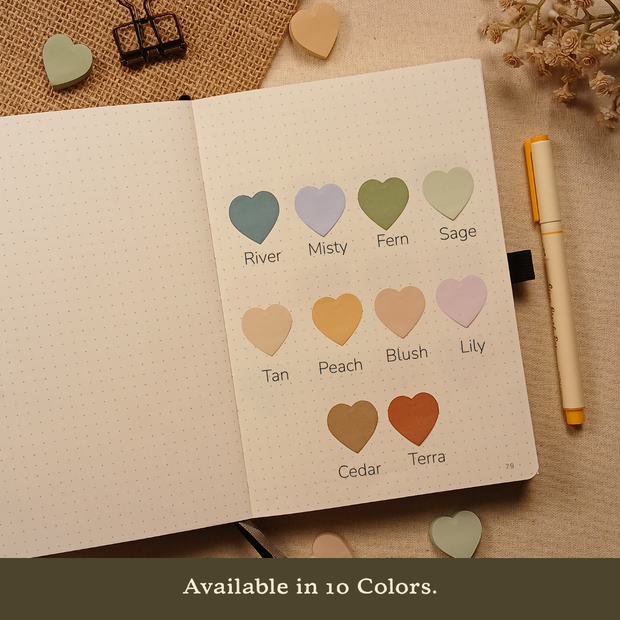 Open bullet journal displaying ten heart-shaped sticky note colors labeled River, Misty, Fern, Sage, Tan, Peach, Blush, Lily, Cedar, and Terra, with the text "Available in 10 Colors."