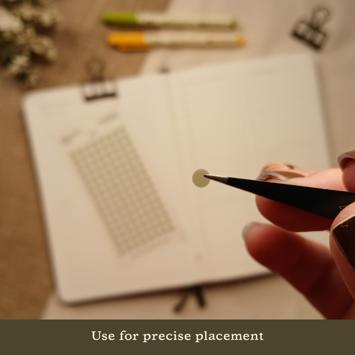 Close-up of black precision tweezers holding a transparent mini dot planner sticker above an open planner, with the text 