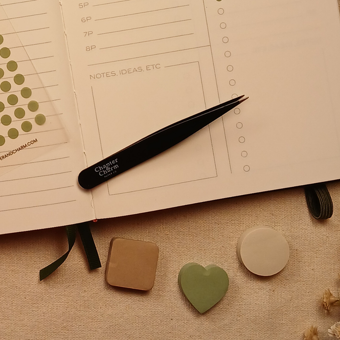 Black precision tweezers resting on an open planner beside heart, circle, and rounded square sticky notes, showing how to easily place planner stickers and sticky notes with accuracy.