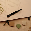 Black precision tweezers resting on an open planner beside heart, circle, and rounded square sticky notes, showing how to easily place planner stickers and sticky notes with accuracy.