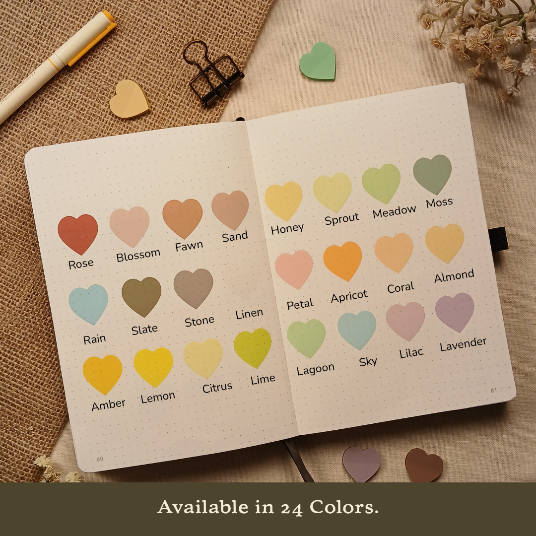 Open bullet journal displaying 24 transparent heart-shaped sticky note colors labeled Rose, Blossom, Fawn, Sand, Honey, Sprout, Meadow, Moss, Rain, Slate, Stone, Linen, Petal, Apricot, Coral, Almond, Amber, Lemon, Citrus, Lime, Lagoon, Sky, Lilac, and Lavender, with the text 