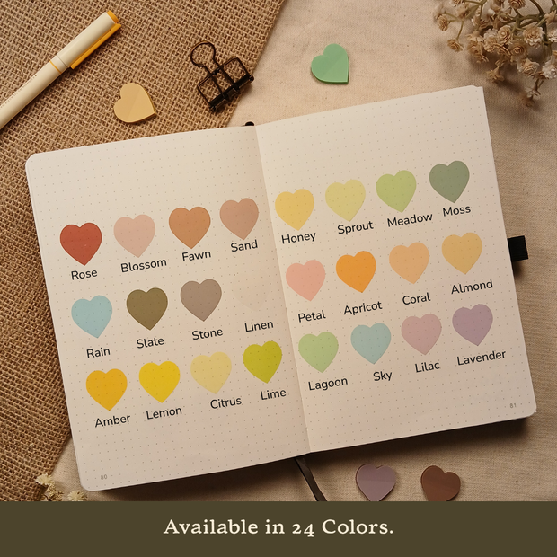 Open bullet journal displaying 24 transparent heart-shaped sticky note colors labeled Rose, Blossom, Fawn, Sand, Honey, Sprout, Meadow, Moss, Rain, Slate, Stone, Linen, Petal, Apricot, Coral, Almond, Amber, Lemon, Citrus, Lime, Lagoon, Sky, Lilac, and Lavender, with the text "Available in 24 Colors."
